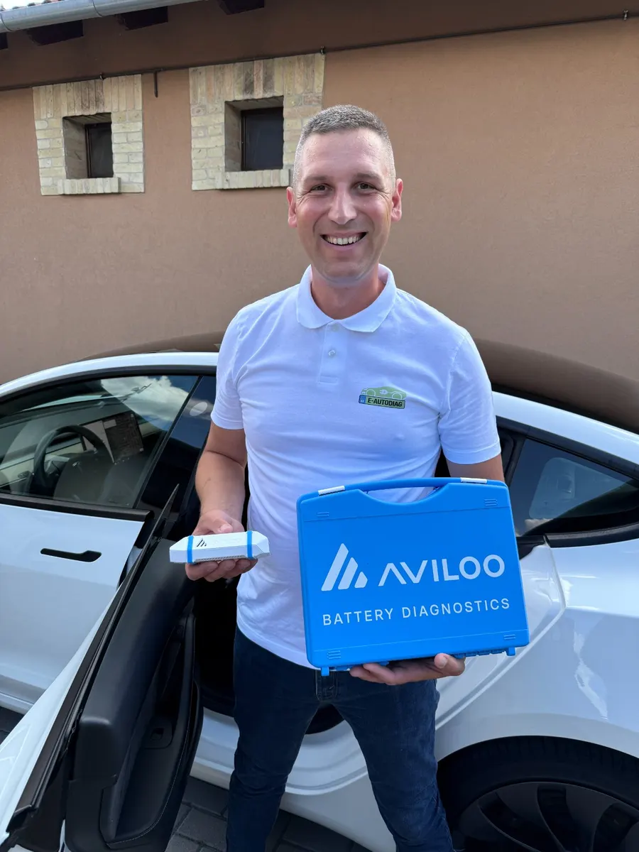 Kapás Krisztián az AVILOO akkumulátor-diagnosztikai eszközzel és kofferrel egy fehér Tesla előtt